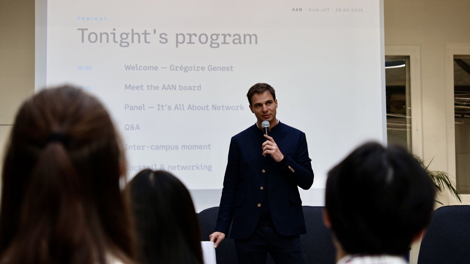 A speaker addressing the room during the kick-off, with the evening programme on screen.